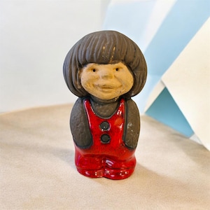 May include: A ceramic figurine of a smiling person with dark hair and a red outfit. The figure has a round face and is wearing a red jumpsuit with black accents. The figurine is standing upright and has a whimsical appearance.