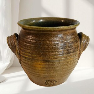 Peut inclure: Un pot en céramique marron de forme ronde avec une large ouverture et deux anses latérales. L'extérieur présente des stries horizontales et une finition émaillée texturée et mouchetée. Une marque de potier circulaire est visible près de la base.