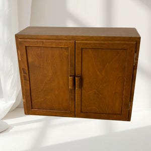 Peut inclure: Une armoire murale rectangulaire en bois avec une finition brun moyen. L'armoire comporte deux portes battantes, chacune dotée d'une simple poignée verticale en bois centrée sur le bord intérieur. Le cadre a un design plat et épuré.
