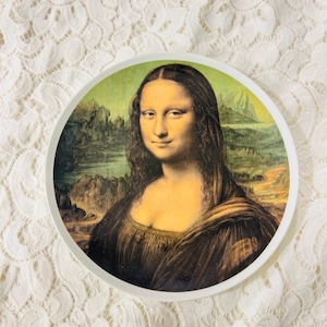 Op de afbeelding: Een ronde decoratieve plaat met het schilderij van de Mona Lisa. De plaat heeft een witte rand en toont het beroemde portret met zijn karakteristieke kleuren en details. De achtergrond van het schilderij toont een landschap.