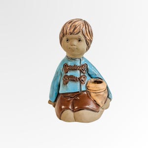 May include: A ceramic figurine of a seated child. The child has light brown hair and is wearing a light blue jacket with brown accents and brown shorts. The child is holding a tan-coloured pot.