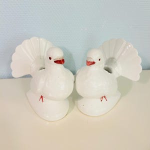 Puede incluir: Dos figuras de palomas de cerámica blanca con picos y patas rojas, y plumas de cola en abanico. Las palomas están posadas sobre una superficie blanca sobre un fondo azul claro. Figuras decorativas de aves.