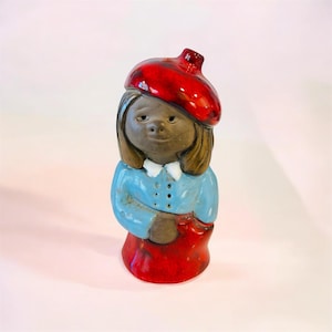 May include: A ceramic figurine of a girl wearing a red beret and a blue dress with a white collar. The figurine has brown hair and a red skirt. The figure is standing upright.
