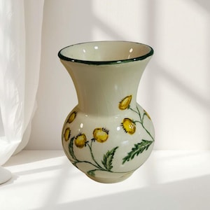 May include: A cream-colored ceramic vase with a flared rim and a rounded body. The vase features a hand-painted botanical design of yellow flowers and green leaves, accented by a thin dark green painted line along the top rim edge.
