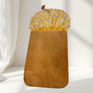May include: A wooden cutting board with a smooth, rectangular base and a rounded top edge. The top section features a carved, three-dimensional relief of wheat stalks painted in shades of yellow, light pink, and blue, with a small brown hanging loop.