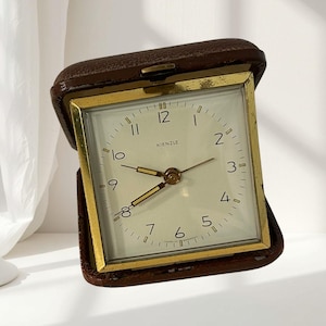 Op de afbeelding: Een vierkante Kienzle reiswekker in een donkerbruine, getextureerde lederen etui. De wijzerplaat is gebroken wit met zwarte Arabische cijfers, goudkleurige wijzers en een goudkleurig metalen frame. De tekst Kienzle staat op de wijzerplaat.