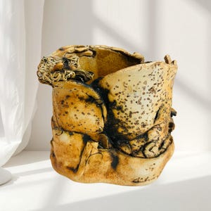 Może przedstawiać: Cylindryczne naczynie ceramiczne o nieregularnej, organicznej krawędzi i teksturowanej powierzchni. Zewnętrzna strona pokryta jest nakrapianym żółto-brązowym szkliwem z ciemnobrązowymi, przypalonymi akcentami oraz drobnymi wypukłymi kropkami.