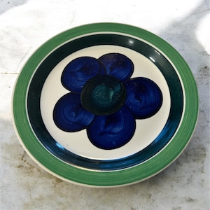 Puede incluir: Un plato redondo de cerámica con un centro blanco con un diseño de flor azul. El plato tiene un borde verde y un anillo azul oscuro. La flor tiene un centro verde azulado oscuro y cinco pétalos azules.