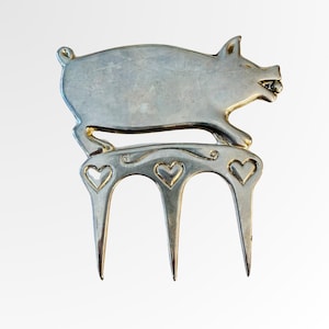Puede incluir: Un tenedor de servir de metal plateado con forma de cerdo. El cerdo se representa a medio paso sobre tres púas. El diseño del tenedor incluye recortes en forma de corazón y adornos decorativos, creando un accesorio de cocina caprichoso.