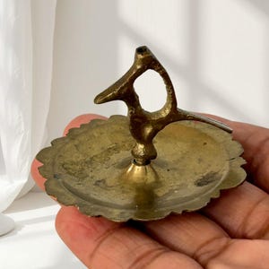 Puede incluir: Un pequeño plato circular de latón con borde festoneado y un asa central vertical con forma de pájaro estilizado. La superficie metálica presenta una pátina envejecida y desgastada con textura visible y manchas de oxidación por toda la pieza.