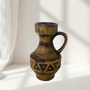 May include: A brown ceramic pitcher with a textured surface, featuring a wide neck with a rectangular pattern and a base decorated with a repeating triangle and dot motif. The vessel has a single curved handle on the right side and a dark finish.