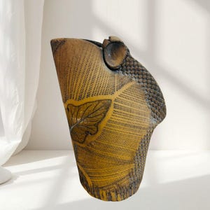 Può includere: Vaso in ceramica fatto a mano con una forma organica unica. Il vaso presenta una superficie strutturata marrone dorato con un motivo a foglie e un motivo a scaglie sul lato. Il vaso è alto circa 30 cm.