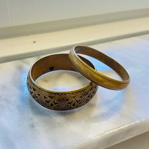 Puede incluir: Dos pulseras doradas. Una pulsera tiene un intrincado diseño calado con un interior oscuro. La otra es una banda lisa y sencilla. Ambas descansan sobre una superficie blanca.