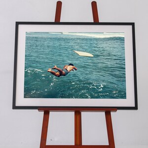 Pu&ograve; includere: Una stampa fotografica incorniciata su un cavalletto in legno che ritrae una persona che si tuffa in un oceano blu profondo. Una tavola da surf chiara galleggia nelle vicinanze sulla superficie del mare. L'opera &egrave; in una cornice nera con passe-partout bianco.