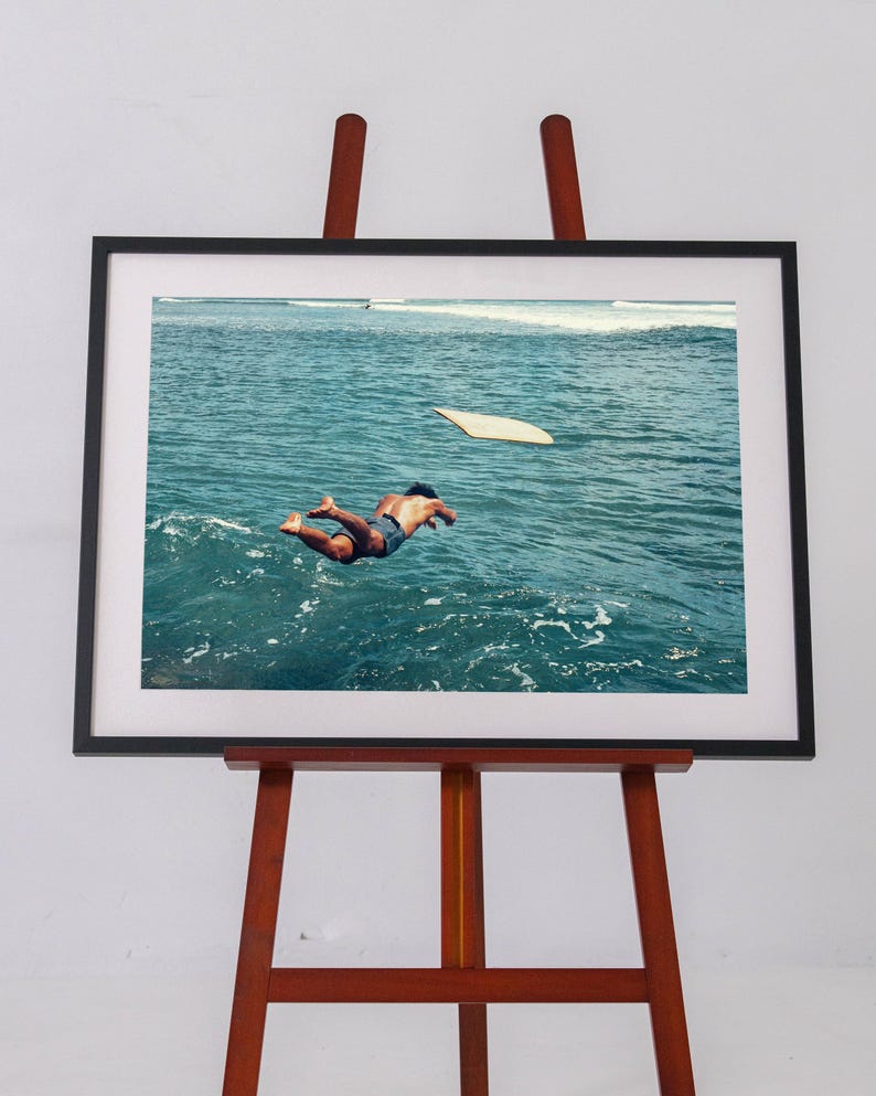 Pu&ograve; includere: Una stampa fotografica incorniciata su un cavalletto in legno che ritrae una persona che si tuffa in un oceano blu profondo. Una tavola da surf chiara galleggia nelle vicinanze sulla superficie del mare. L'opera &egrave; in una cornice nera con passe-partout bianco.