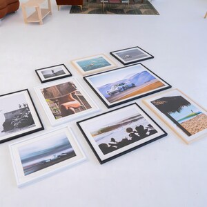 Pu&ograve; includere: Una collezione di dieci stampe fotografiche incorniciate disposte su un pavimento bianco. Le cornici sono nere o in legno chiaro e contengono varie immagini, tra cui un'auto da corsa, una scena di spiaggia, una silhouette e paesaggi costieri.