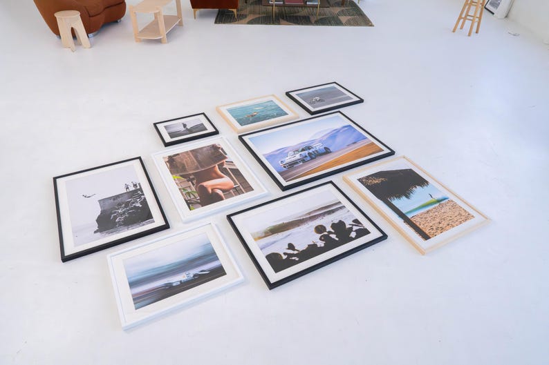 Pu&ograve; includere: Una collezione di dieci stampe fotografiche incorniciate disposte su un pavimento bianco. Le cornici sono nere o in legno chiaro e contengono varie immagini, tra cui un'auto da corsa, una scena di spiaggia, una silhouette e paesaggi costieri.