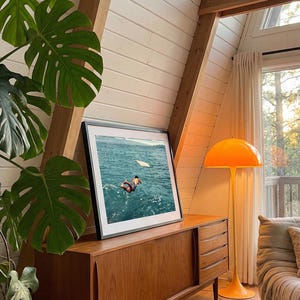 Pu&ograve; includere: Una fotografia incorniciata di una persona che si tuffa nell'acqua blu dell'oceano, appoggiata su una credenza in legno. La credenza presenta ante scorrevoli e cassetti. Accanto si trova una lampada da terra arancione a forma di fungo.