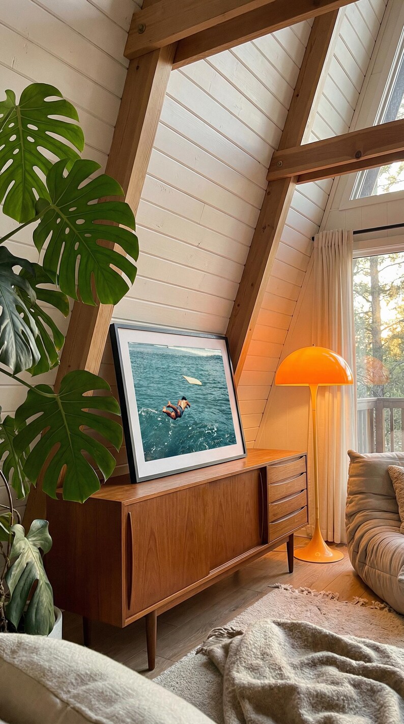 Pu&ograve; includere: Una fotografia incorniciata di una persona che si tuffa nell'acqua blu dell'oceano, appoggiata su una credenza in legno. La credenza presenta ante scorrevoli e cassetti. Accanto si trova una lampada da terra arancione a forma di fungo.