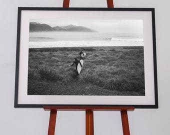 May include: A black and white framed photograph displayed on a wooden easel. The image depicts two individuals carrying surfboards through tall beach grass toward the ocean, with distant mountains visible under a cloudy sky in the background.
