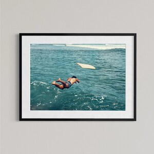 Pu&ograve; includere: Una stampa artistica incorniciata che ritrae una persona che si tuffa in acque oceaniche blu intenso con una tavola da surf chiara che galleggia nelle vicinanze. La scena cattura un momento di movimento sopra le onde in una cornice nera.
