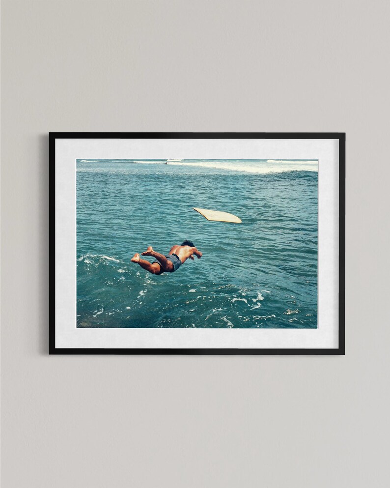 Pu&ograve; includere: Una stampa artistica incorniciata che ritrae una persona che si tuffa in acque oceaniche blu intenso con una tavola da surf chiara che galleggia nelle vicinanze. La scena cattura un momento di movimento sopra le onde in una cornice nera.