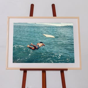 Pu&ograve; includere: Una stampa fotografica incorniciata su un cavalletto in legno, che ritrae una persona che si tuffa in acque oceaniche blu intenso. Una tavola da surf chiara galleggia nelle vicinanze sulla superficie del mare, con schiuma bianca in lontananza.