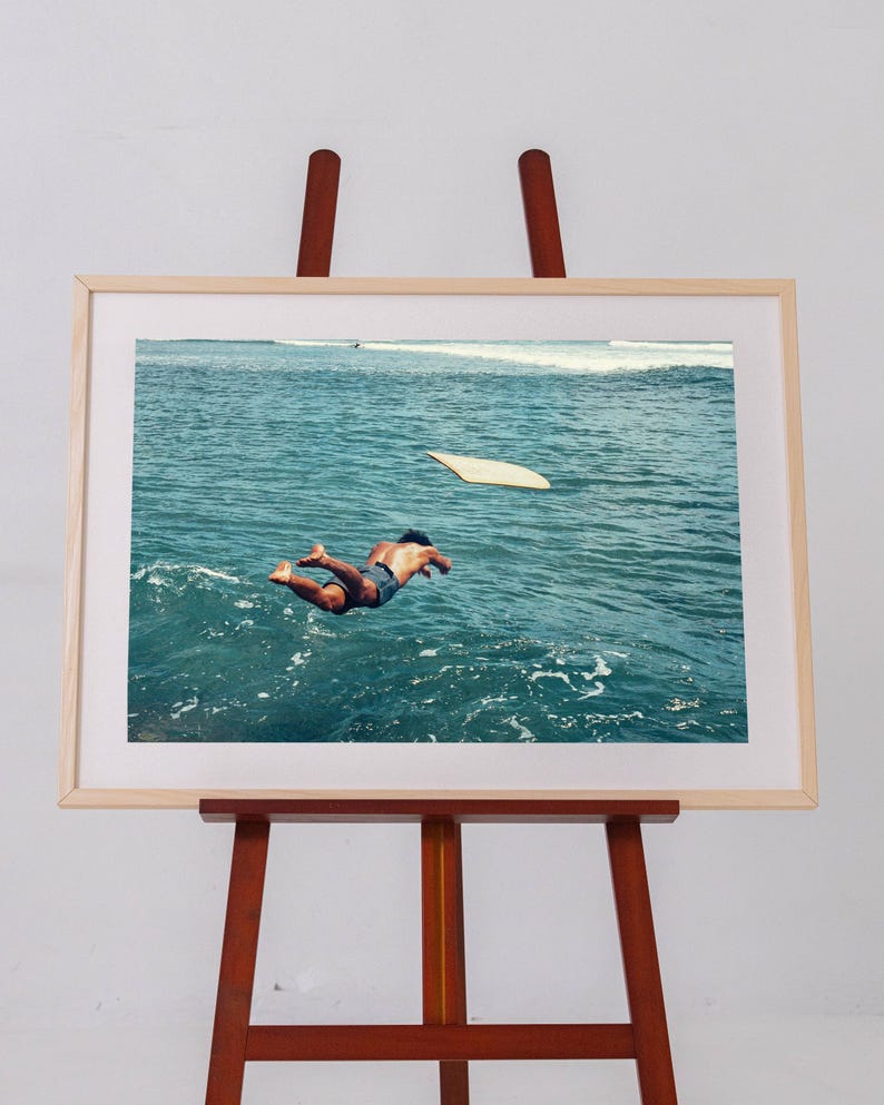 Pu&ograve; includere: Una stampa fotografica incorniciata su un cavalletto in legno, che ritrae una persona che si tuffa in acque oceaniche blu intenso. Una tavola da surf chiara galleggia nelle vicinanze sulla superficie del mare, con schiuma bianca in lontananza.
