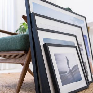 Pu&ograve; includere: Un set di quattro cornici nere annidate con passepartout bianco, ognuna contenente una diversa stampa fotografica. Le cornici sono disposte a cascata e mostrano varie immagini costiere e oceaniche, tra cui un surfista su un'onda.