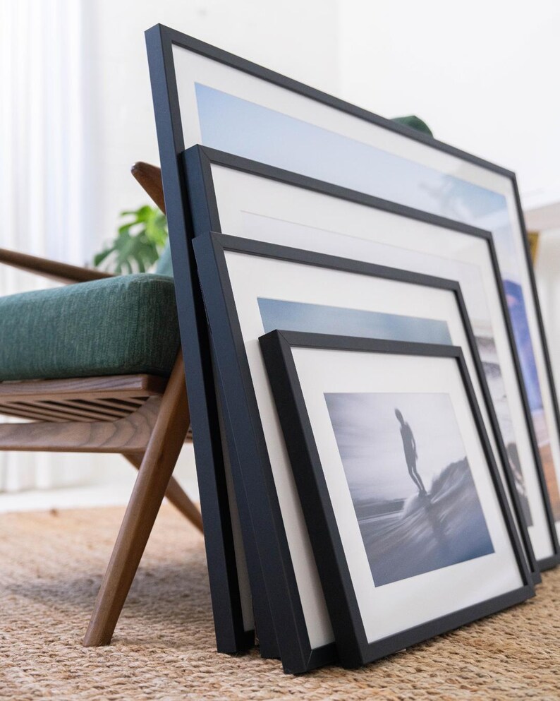 Pu&ograve; includere: Un set di quattro cornici nere annidate con passepartout bianco, ognuna contenente una diversa stampa fotografica. Le cornici sono disposte a cascata e mostrano varie immagini costiere e oceaniche, tra cui un surfista su un'onda.