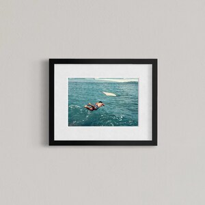 Pu&ograve; includere: Un'opera d'arte da parete incorniciata che presenta una fotografia di una persona che nuota in acque oceaniche blu intenso verso una tavola da surf bianca. L'opera &egrave; racchiusa in una semplice cornice rettangolare nera con un ampio passe-partout.