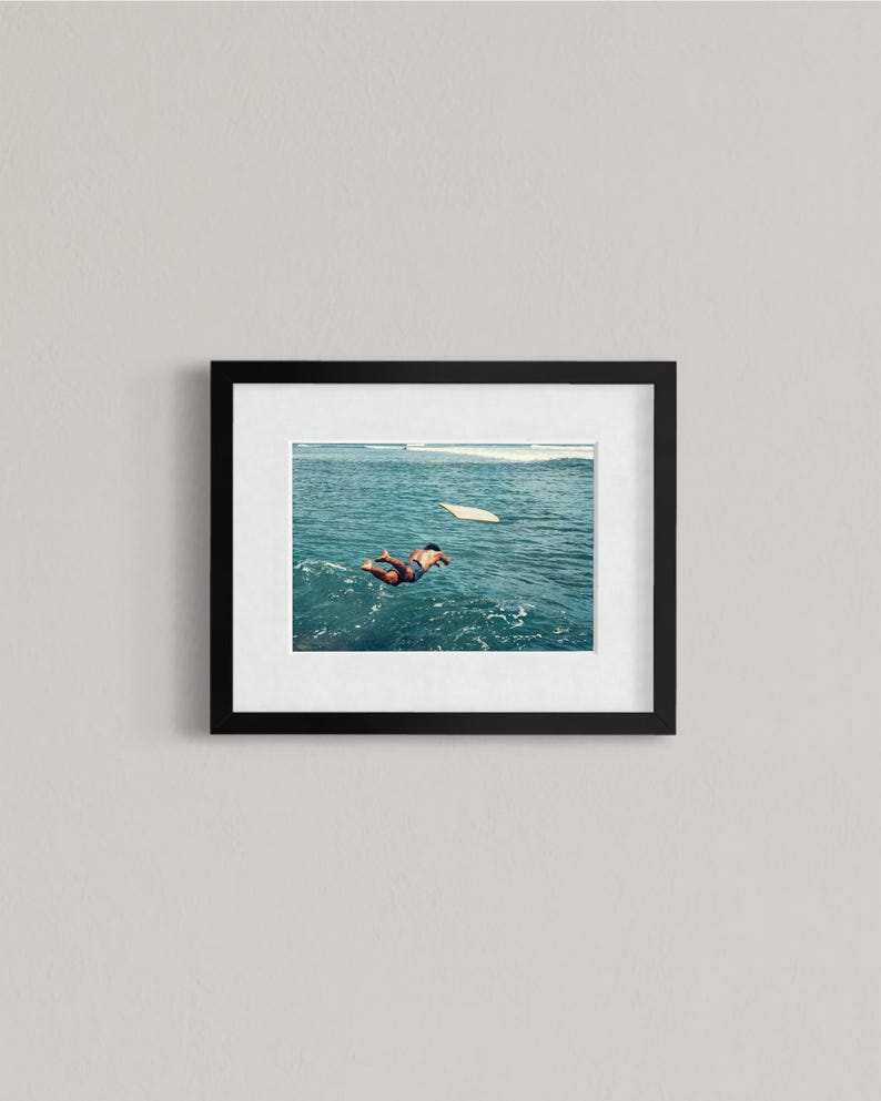 Pu&ograve; includere: Un'opera d'arte da parete incorniciata che presenta una fotografia di una persona che nuota in acque oceaniche blu intenso verso una tavola da surf bianca. L'opera &egrave; racchiusa in una semplice cornice rettangolare nera con un ampio passe-partout.