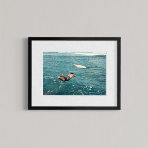 Pu&ograve; includere: Una stampa artistica incorniciata che mostra la fotografia di una persona che si tuffa in acque oceaniche blu intenso, con una tavola da surf bianca che galleggia nelle vicinanze. L'opera &egrave; racchiusa in una cornice nera con passe-partout bianco.