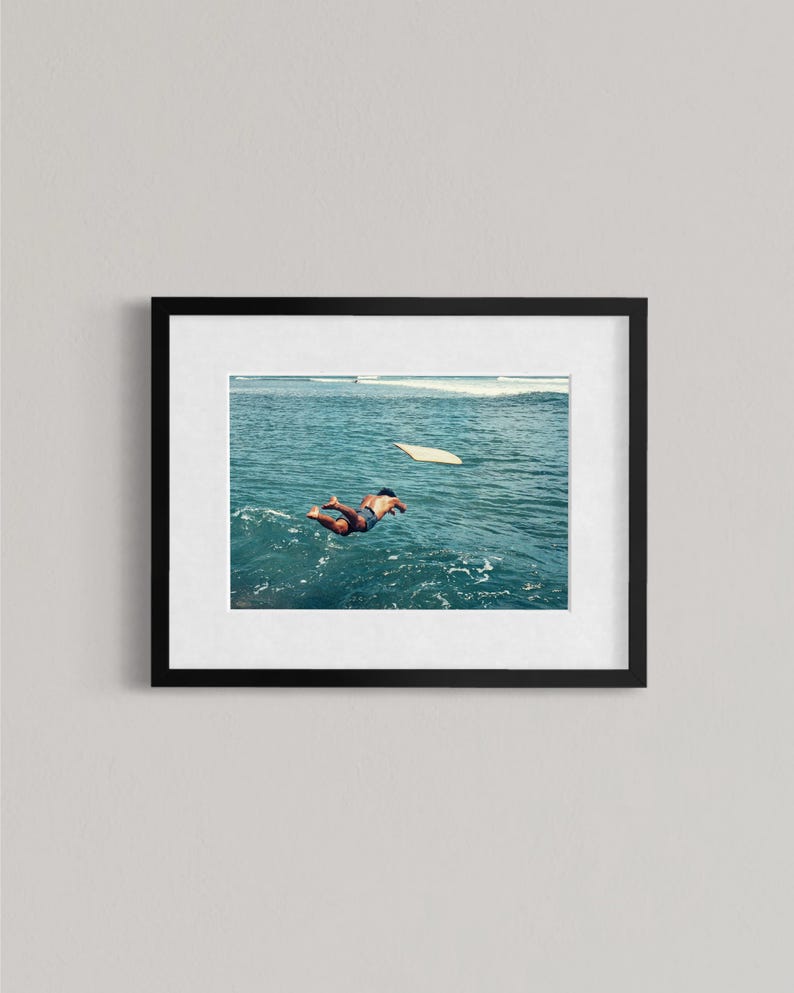 Pu&ograve; includere: Una stampa artistica incorniciata che mostra la fotografia di una persona che si tuffa in acque oceaniche blu intenso, con una tavola da surf bianca che galleggia nelle vicinanze. L'opera &egrave; racchiusa in una cornice nera con passe-partout bianco.