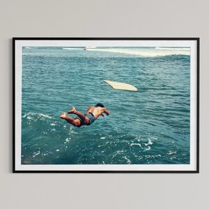 Pu&ograve; includere: Una stampa artistica incorniciata che ritrae una persona che si tuffa in acque oceaniche blu intenso. Una tavola da surf di colore chiaro galleggia in superficie nelle vicinanze. La scena cattura il movimento del tuffo tra le onde del mare.