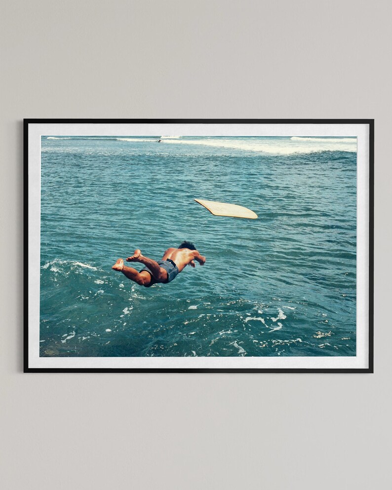 Pu&ograve; includere: Una stampa artistica incorniciata che ritrae una persona che si tuffa in acque oceaniche blu intenso. Una tavola da surf di colore chiaro galleggia in superficie nelle vicinanze. La scena cattura il movimento del tuffo tra le onde del mare.