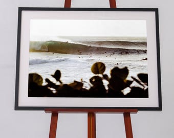 Può includere: Una fotografia paesaggistica incorniciata di un'onda che si infrange con diversi surfisti in acqua, esposta su un cavalletto di legno. Il primo piano presenta sagome scure e sfocate di foglie tropicali che contrastano con il mare luminoso.