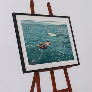 Pu&ograve; includere: Una stampa fotografica incorniciata esposta su un cavalletto in legno. L'immagine ritrae una persona che si tuffa in acque oceaniche blu intenso, con una tavola da surf chiara che galleggia nelle vicinanze. La cornice &egrave; nera con un ampio passe-partout bianco.