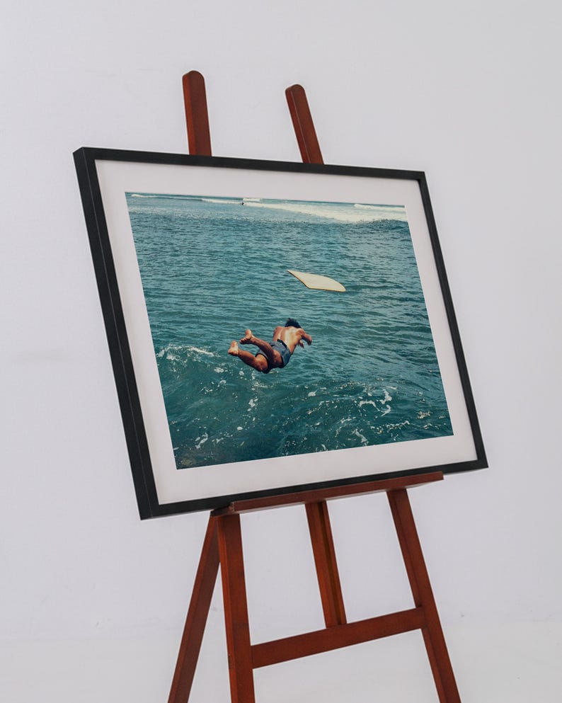 Pu&ograve; includere: Una stampa fotografica incorniciata esposta su un cavalletto in legno. L'immagine ritrae una persona che si tuffa in acque oceaniche blu intenso, con una tavola da surf chiara che galleggia nelle vicinanze. La cornice &egrave; nera con un ampio passe-partout bianco.