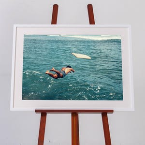 Pu&ograve; includere: Una stampa fotografica incorniciata su un cavalletto in legno, che mostra una persona che si tuffa nell'acqua blu profonda dell'oceano verso una tavola da surf chiara che galleggia in superficie, con onde visibili in lontananza sotto un cielo luminoso.