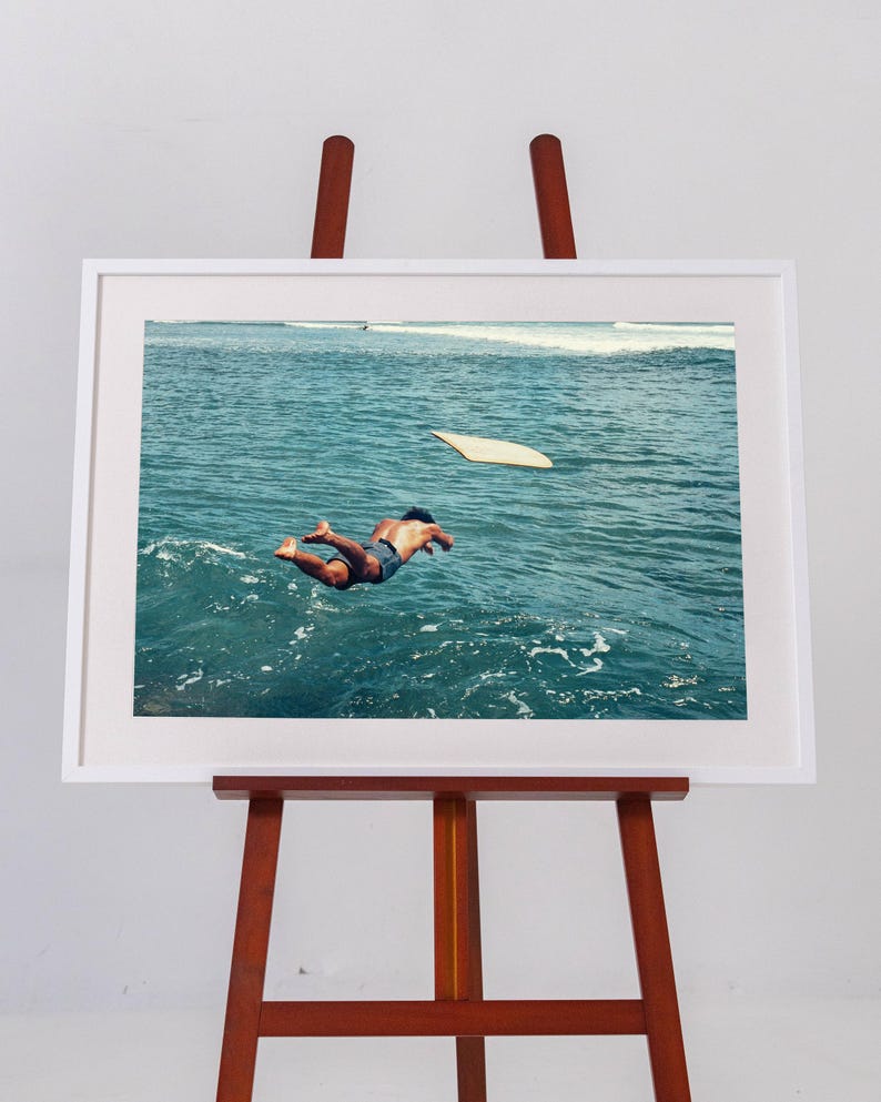 Pu&ograve; includere: Una stampa fotografica incorniciata su un cavalletto in legno, che mostra una persona che si tuffa nell'acqua blu profonda dell'oceano verso una tavola da surf chiara che galleggia in superficie, con onde visibili in lontananza sotto un cielo luminoso.