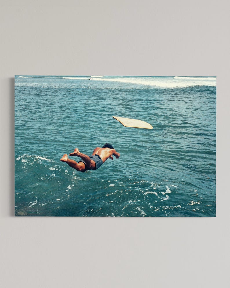 Pu&ograve; includere: Una persona che indossa pantaloncini blu scuro si tuffa nell'acqua blu profondo dell'oceano. Una tavola da surf rettangolare di colore chiaro fluttua nell'aria sopra la superficie dell'acqua, con onde visibili sullo sfondo sotto un cielo limpido.