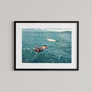 Pu&ograve; includere: Una stampa artistica da parete incorniciata che mostra la fotografia di una persona che si tuffa in acque oceaniche blu intenso. Una tavola da surf galleggia nelle vicinanze sulla superficie del mare, mentre le onde sono visibili in lontananza.