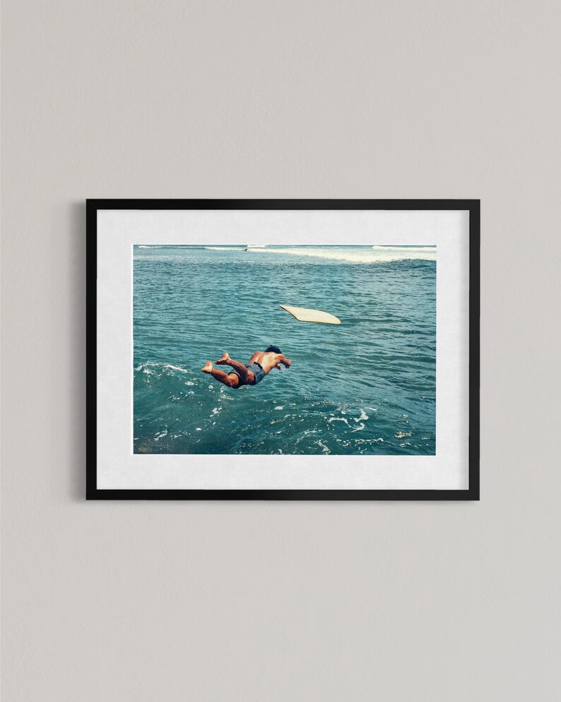 Pu&ograve; includere: Una stampa artistica da parete incorniciata che mostra la fotografia di una persona che si tuffa in acque oceaniche blu intenso. Una tavola da surf galleggia nelle vicinanze sulla superficie del mare, mentre le onde sono visibili in lontananza.