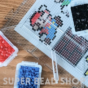 May include: A close-up of a clear plastic bead board with a pixelated image of a video game character. The image is labeled "Super Mario Brother, PAGE 1". There are four small containers of colorful beads, including red, blue, and black.