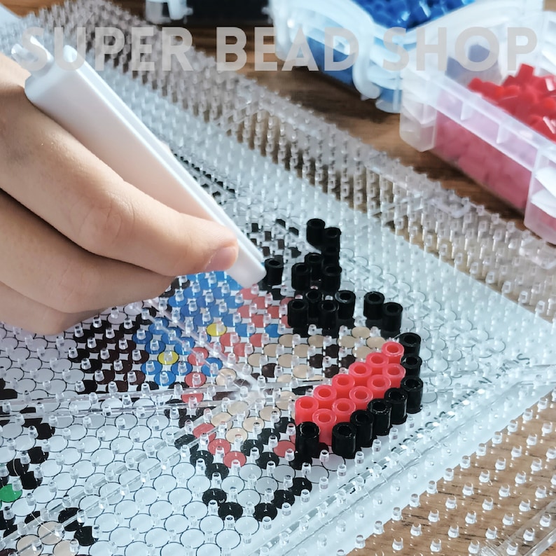 May include: A close-up of a clear plastic bead board with red, black, and white beads arranged in a pattern. A white tool is being used to press the beads into place.