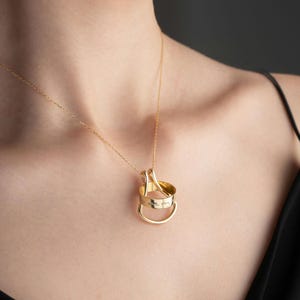 May include: A gold pendant necklace with a three-ring design. The rings are linked and hang from a fine gold chain. The bottom ring has the inscription "love my". The necklace is displayed against a dark backdrop.