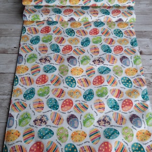 May include: A white Easter-themed fabric with a repeating pattern of colorful Easter eggs. The eggs feature various designs, including stripes, floral patterns, and text that reads "Easter" and "A happy Easter". The fabric is rolled up.