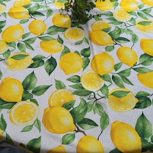 Può includere: Una tovaglia rotonda con un motivo a limoni vivace. Il tessuto bianco è decorato con limoni gialli, alcuni tagliati a metà, e foglie verdi. Una piccola pianta in vaso è posta al centro del tavolo, aggiungendo un tocco di verde alla scena.