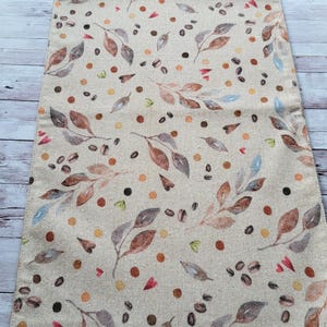 Può includere: Runner da tavolo beige con un motivo ripetuto di foglie, chicchi di caffè e piccoli puntini nei toni del marrone, rosso e verde. Il runner è realizzato in tessuto strutturato ed è arrotolato nella parte superiore.